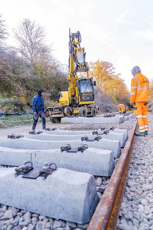 Concrete ties are laid with an excavator along a railway line, supervised by workers, track construction of the Hermann Hesse Railway, Calw, Germany
