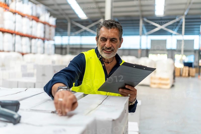 Logistics manager smiling while using a barcode scanner and clipboard checking shipment boxes in a large modern distribution warehouse, ensuring efficient supply chain operations
