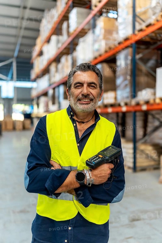 Middle aged man wearing safety vest and holding a barcode scanner, smiling, while standing with arms crossed in a large logistics warehouse with shelves of products