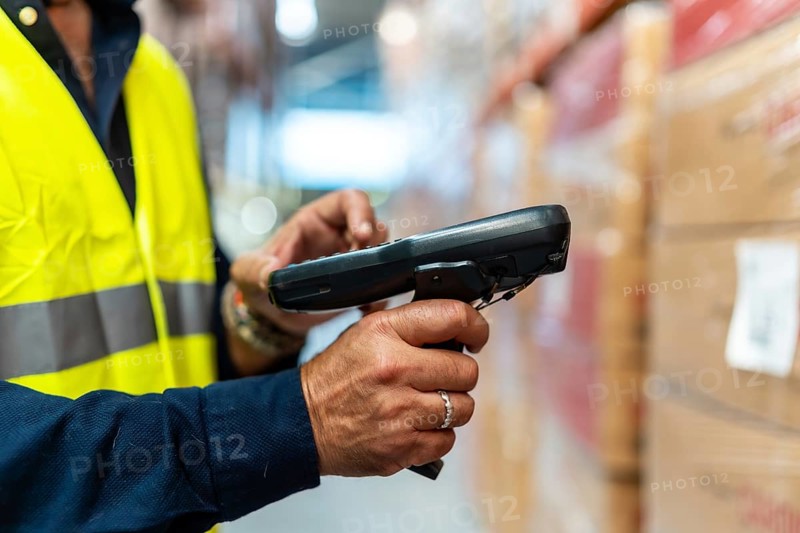 Warehouse worker in high visibility vest and blue uniform scanning barcodes on boxes with handheld scanner, updating inventory and tracking stock in a busy distribution center