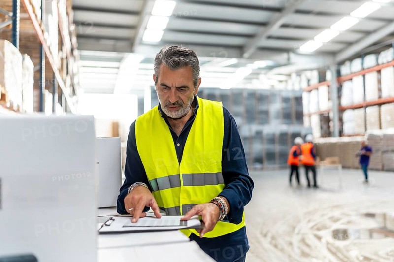 Experienced manager wearing a high visibility safety vest confirming details on a clipboard while standing in a busy logistics warehouse, overseeing stock and supply chain operations