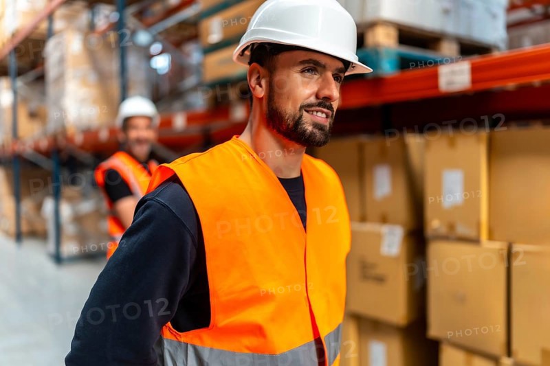 Man wearing an orange safety vest and white hard hat, working at a logistics warehouse with shelves stacked high with boxes, ensuring efficient operations