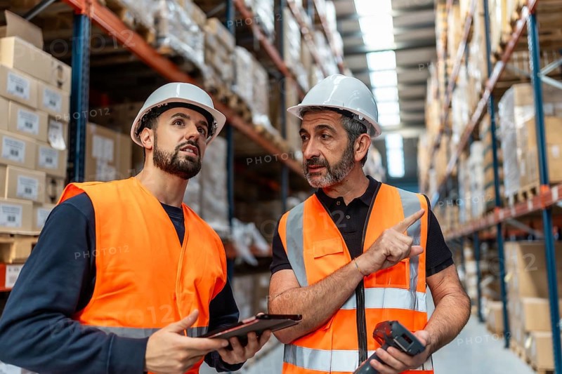 Two male warehouse workers wearing safety vests and hard hats discussing inventory with a digital tablet and barcode scanner, standing between tall racks of stacked boxes