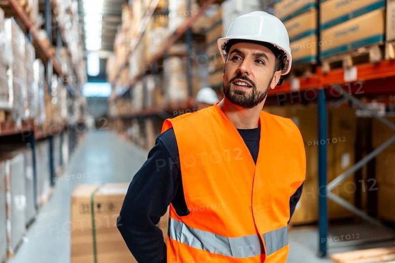 Warehouse worker in hard hat and orange safety vest standing in an aisle, inspecting high shelves of inventory and boxes, checking stock in a modern distribution facility