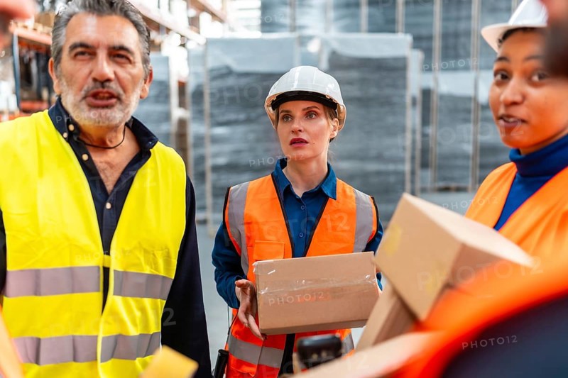 Diverse group of logistics professionals wearing safety vests and hard hats discussing shipping operations and inventory management in a modern distribution warehouse