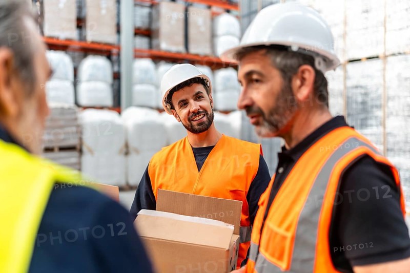 Warehouse workers wearing safety vests and hard hats are discussing tasks while one man holds a cardboard box in a bustling logistics facility with shelves full of products