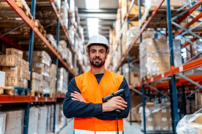 Male logistics worker in safety helmet and uniform smiles with crossed arms and a handheld barcode scanner in a busy distribution warehouse, representing efficient shipping and inventory