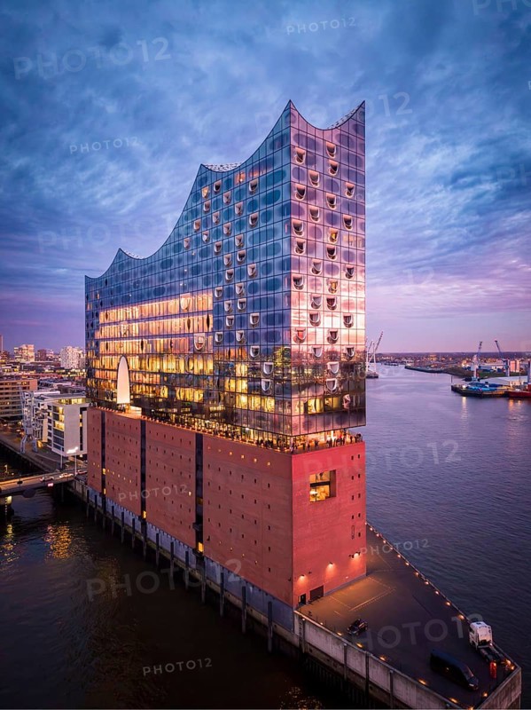 Aerial view of the Elbe Philharmonic Hall with reflections on the façade in evening light above the harbor, surrounded by water and urban backdrop, Hamburg, Germany