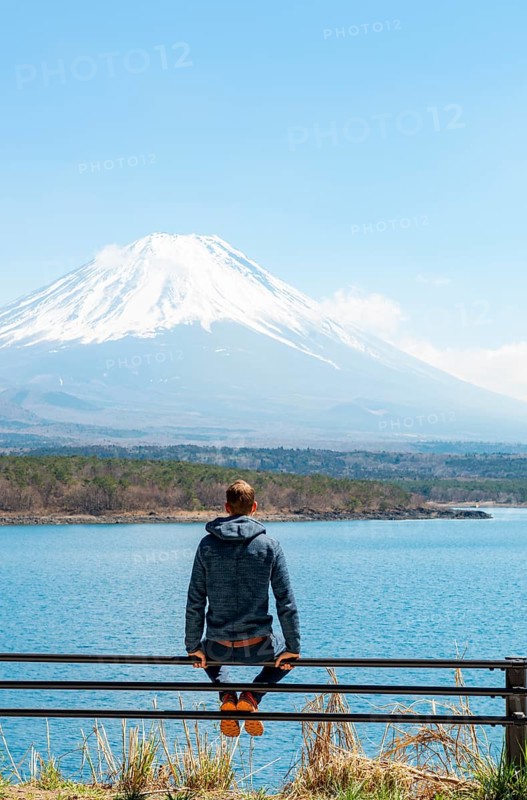 Young man sitting on railings next to a road and looking across the lake to Mt Fuji volcano, Motosu Lake, Yamanashi Prefecture, Japan