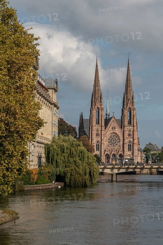 The two-towered church stands majestically on the banks of the river. Trees with golden leaves surround historic buildings under cloudless ski slopes. Bas Rhin, Alsace, France