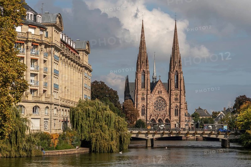 The two-towered church stands majestically on the banks of the river. Trees with golden leaves surround historic buildings under cloudless ski slopes. Bas rhin, Alsace, Grand Est, France