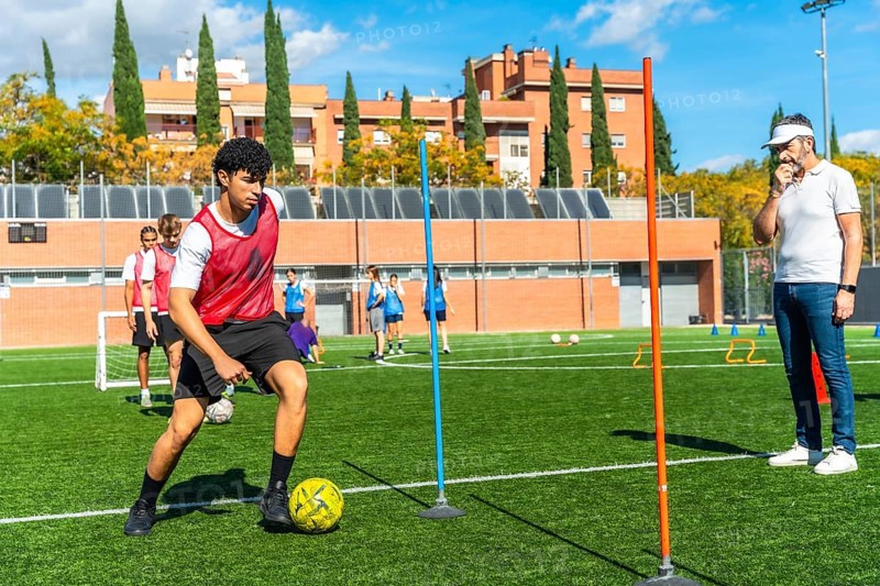 Young male student athlete concentrating on dribbling a soccer ball between poles during a drills practice session, with a coach observing and other players in the background