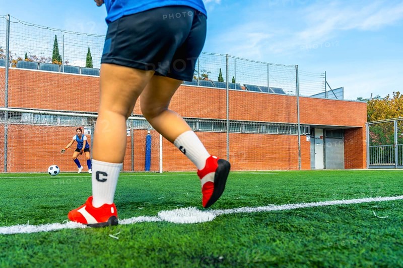 Football player preparing to shoot a penalty kick on an artificial turf field, with the opposing team's goalkeeper ready to defend the goal in the background