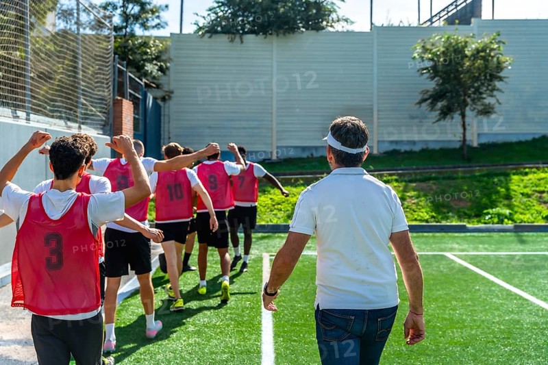 Youth soccer coach leading a group of players in red training vests across artificial grass, preparing the team for practice with guidance, teamwork and focus
