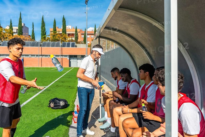 Soccer coach distributing water bottles to young male players resting in the dugout during a training session or match, emphasizing hydration and teamwork