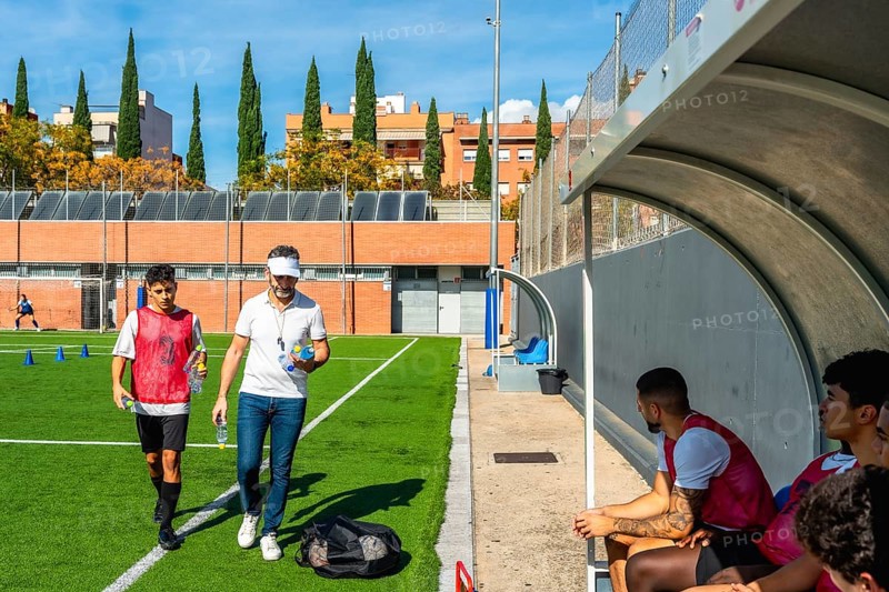 Soccer coach and young player walking on a green artificial turf field after training, carrying water bottles, with other team members resting in the dugout