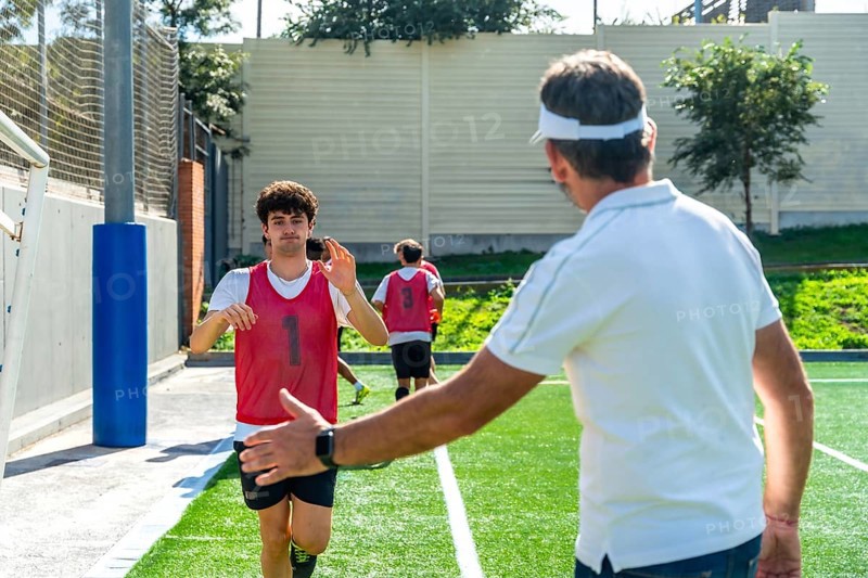 Soccer coach extending hand for a high five with a young male player wearing a red bib during a team training session on a green artificial turf field, with other players in background