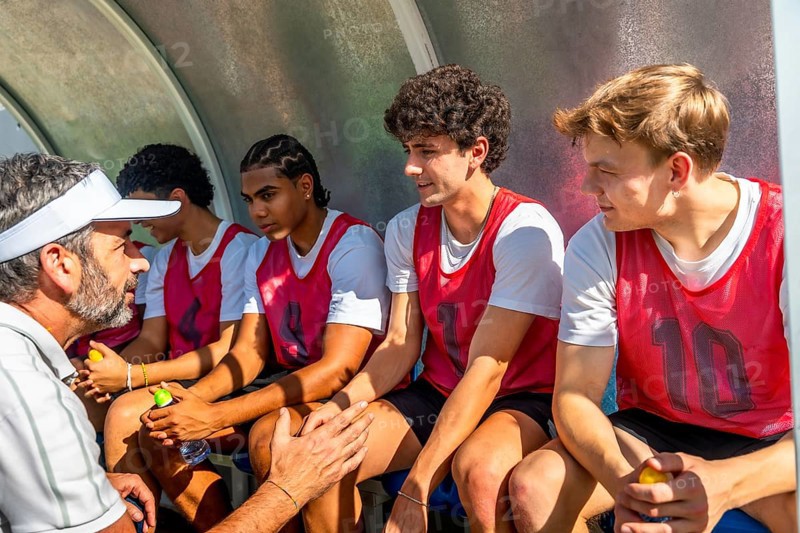Experienced coach providing advice, guidance, and motivation to a group of young diverse male soccer players sitting in the team dugout during a training session