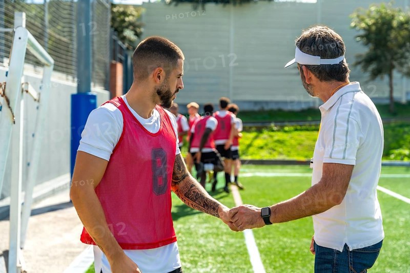 Coach and young soccer player shaking hands on a green sports field, demonstrating teamwork, respect, and sportsmanship during training with other teammates in the background