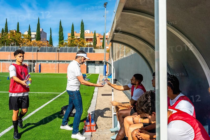 Soccer coach distributing water bottles to young male players sitting in the dugout during a hydration break on the field after an intensive training session
