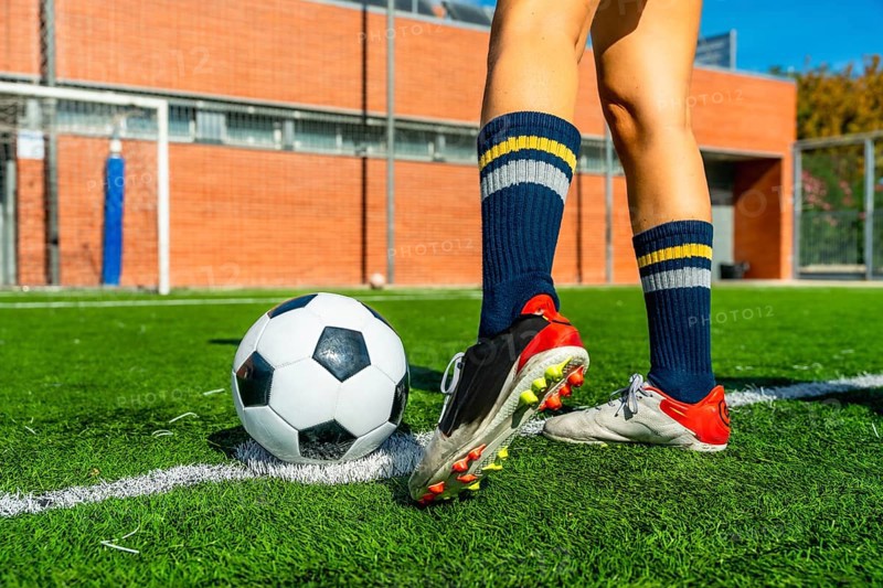 Female soccer player's legs and feet are positioned near a soccer ball on a green artificial turf field with white lines, featuring athletic performance and preparation