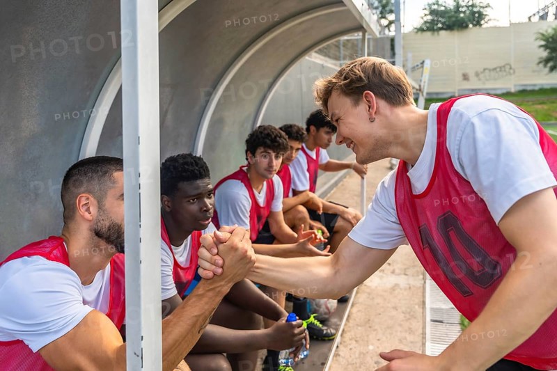 Diverse group of young adult soccer players showing camaraderie and good sportsmanship, shaking hands while sitting on the team bench during a break or before a match