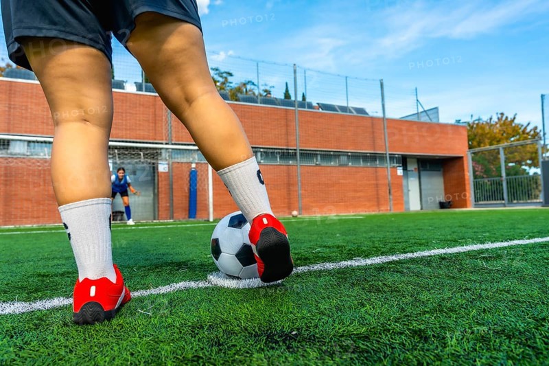 Woman soccer player preparing to strike a penalty kick on a green artificial turf field, with a blurred goalkeeper in the background ready to defend the goal