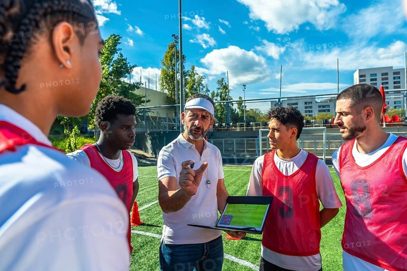 Soccer coach explaining game strategy to a diverse group of male players on a green artificial turf field, showing teamwork, coaching, and a professional plan during training