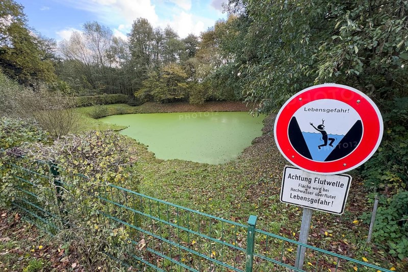 Fence with warning sign with lettering Danger to life and symbolic depiction of person falling into water Reservoir with smooth walls, including warning Attention Futwelle, area will be flooded during floods, danger to life, retention basin filled with water on water surface closed green algae layer, Germany