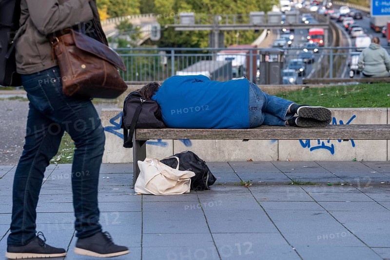 Man sleeping on a bench, travelling bag serves as pillow, in the city centre, passers-by walk by