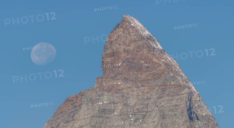 The Matterhorn mountain peak rises steeply against a bright blue sky. The full moon glistens gently above, creating a marvellous view in the serene evening hours. Matterhorn, Zermatt, Valais, Alps, Swiss