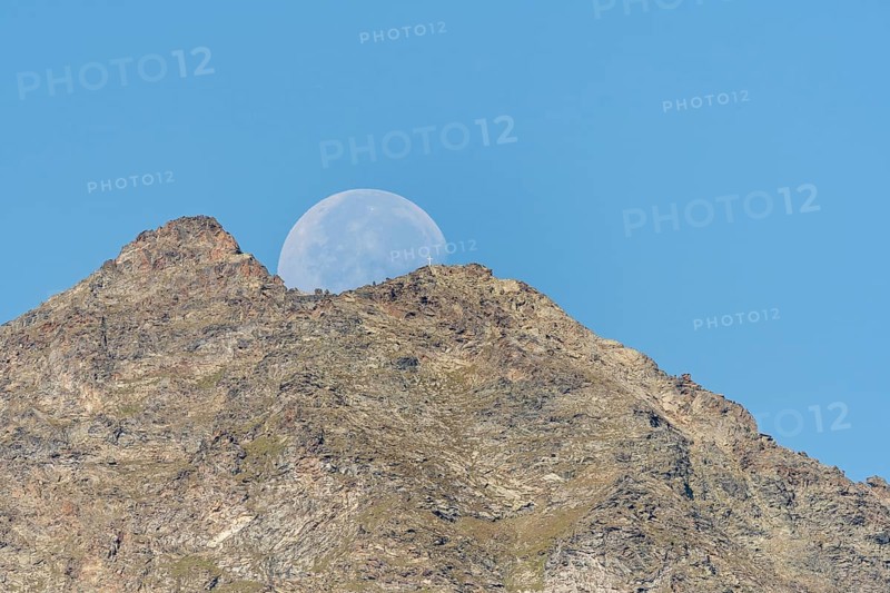 Majestic mountain peak stands tall against vibrant blue sky as large moon rises just behind it. The scene captures the quiet beauty of nature in the early evening light. Grachen, Viege, Valais, Switzerland
