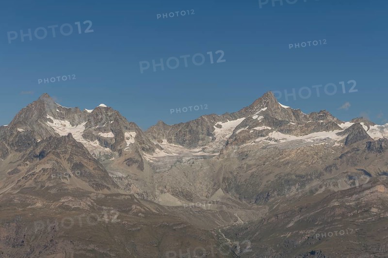 Majestic mountain peak rises prominently against a vibrant blue sky, surrounded by rocky terrain and snow-covered slopes. The sun illuminates the landscape as the clouds drift nearby. Zermatt, Valais, Alps, Swiss