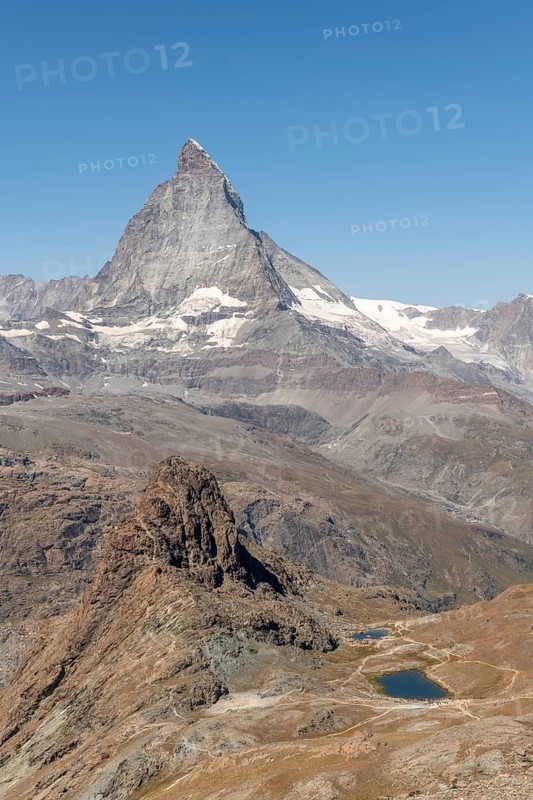 The towering peak of the Matterhorn dominates the clear sky, surrounded by rocky terrain and patches of snow. The barren landscape reveals the natural beauty of the Swiss Alps. Zermatt, Valais