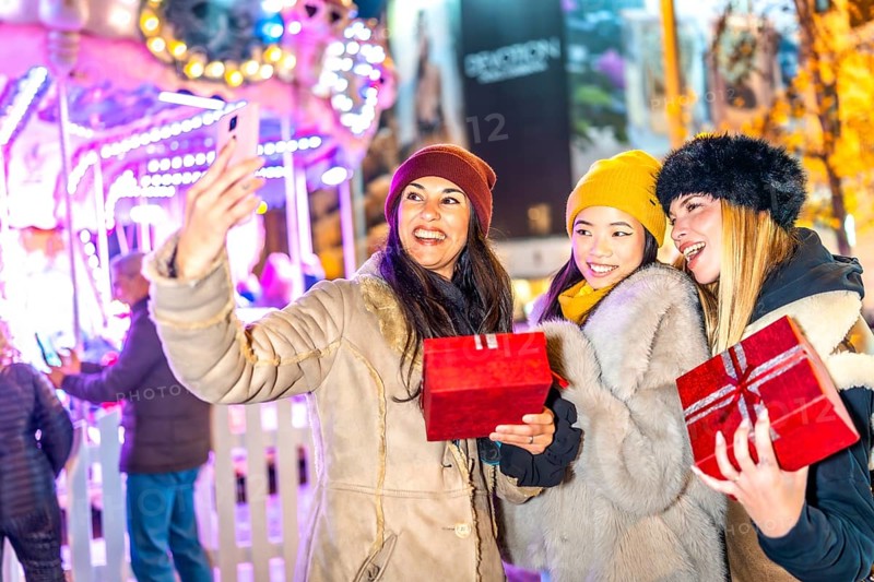 Three happy women holding christmas gifts are taking a selfie in front of a carousel in a christmas market at night
