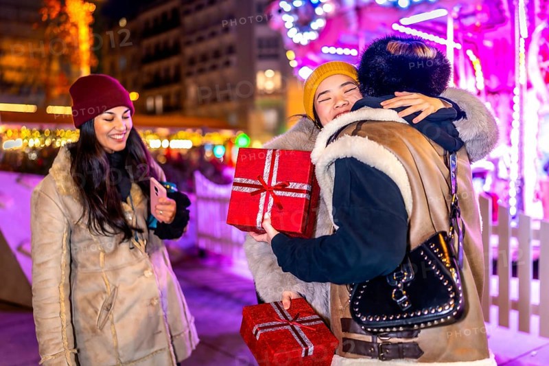 Three happy female friends exchanging christmas gifts and hugging at a christmas market at night