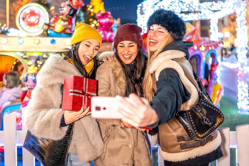 Three happy female friends taking a selfie with a smartphone at a christmas market amusement park, holding christmas gifts