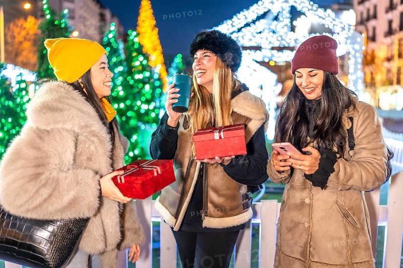 Three cheerful women enjoying christmas night in a decorated city square, holding gifts, drinking hot beverages, and using a smartphone