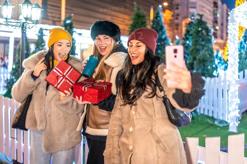 Three cheerful female friends holding christmas gifts and taking a selfie at a decorated outdoor christmas market at night