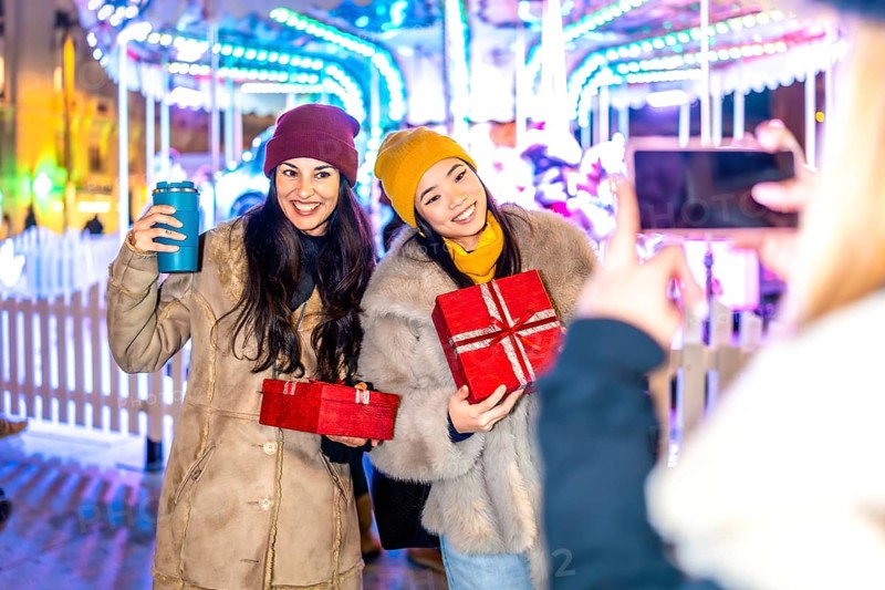 Two happy women holding christmas gifts posing for a picture in front of a carousel at a christmas market, enjoying their winter holidays