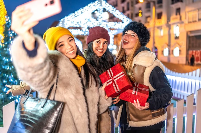 Three cheerful women holding christmas gifts taking a selfie with smartphone at christmas market in the city at night