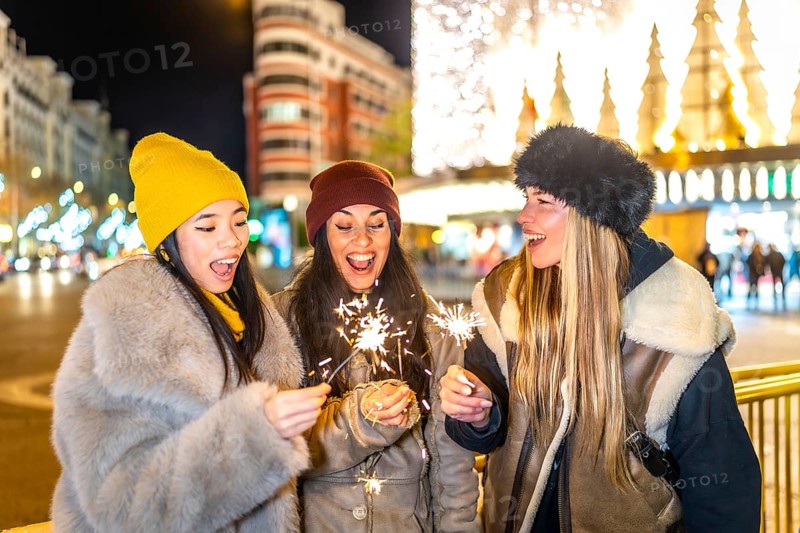 Three happy young women holding burning sparklers, enjoying christmas night in a brightly illuminated city