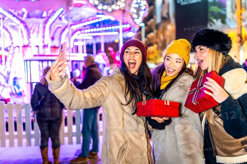 Three cheerful women holding christmas gifts taking a selfie in front of a carousel at night in a christmas themed amusement park