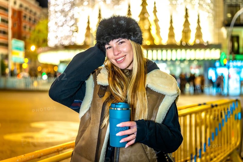 Stylish woman holding reusable cup with hot drink, enjoying magical christmas lights and festive atmosphere in a city at night