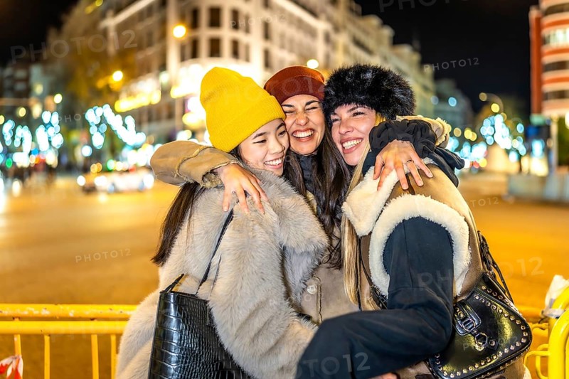 Three cheerful young women hugging each other in a city street decorated with christmas lights