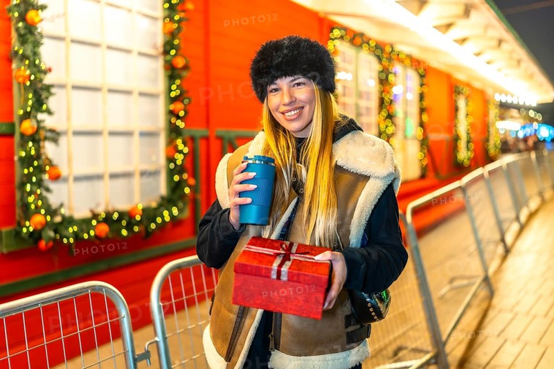 Young woman enjoying a warm beverage and holding a christmas present at a festive night market during the holiday season