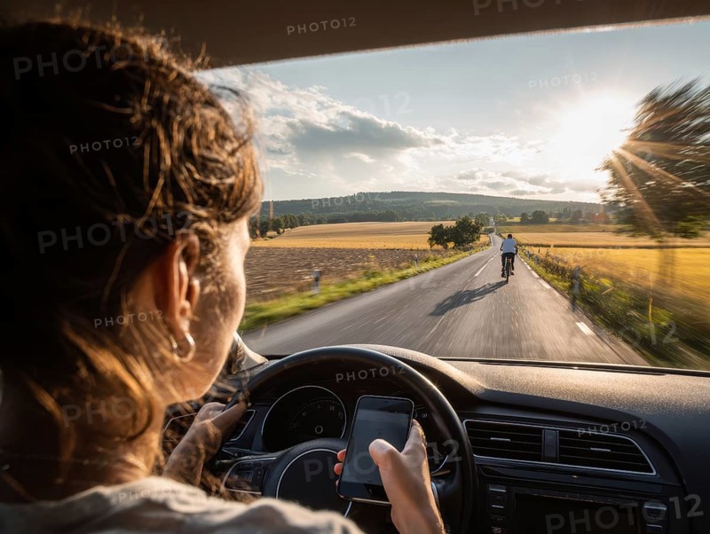 A car driver is dazzled by the sun and looks at her smartphone while driving, putting herself, the cyclist in front and oncoming traffic in danger. Carelessness, recklessness, distraction in road traffic, accident risk, AI generated, AI generated