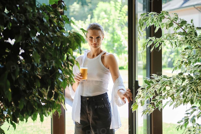 A young woman stands at the entrance of a bright garden space, holding a glass of iced beverage. Dressed in a casual white top and jeans, she appears relaxed amid lush green plants in the background