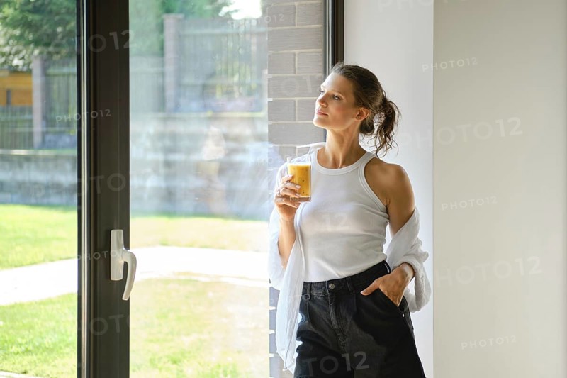 Cute woman stands thoughtfully by a large window, sipping a smoothies in a light-filled space. She wears a casual outfit and displays a relaxed attitude, enjoying the sunny view outdoors