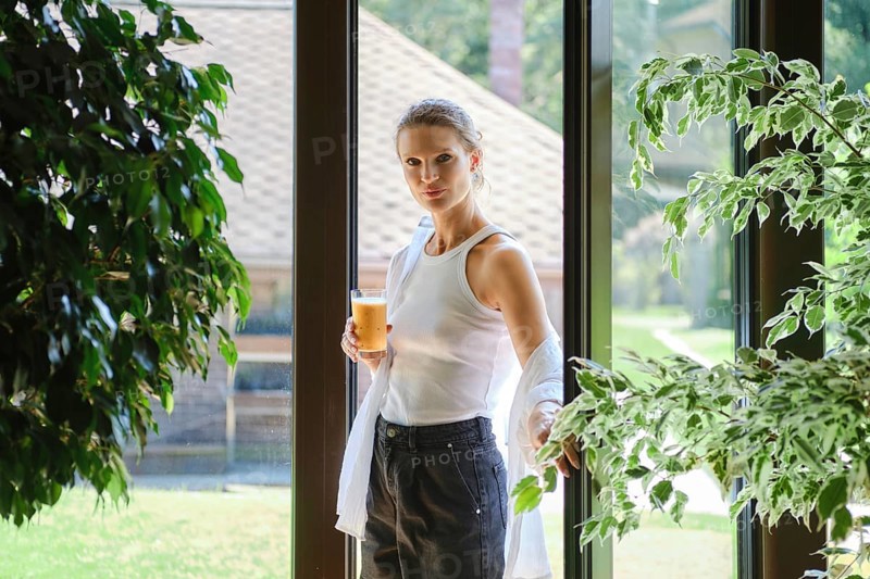 A woman stands by a large window, sipping a cool beverage while surrounded by green plants and sunlight. Outside, a tranquil garden offers a peaceful backdrop, enhancing the relaxing atmosphere